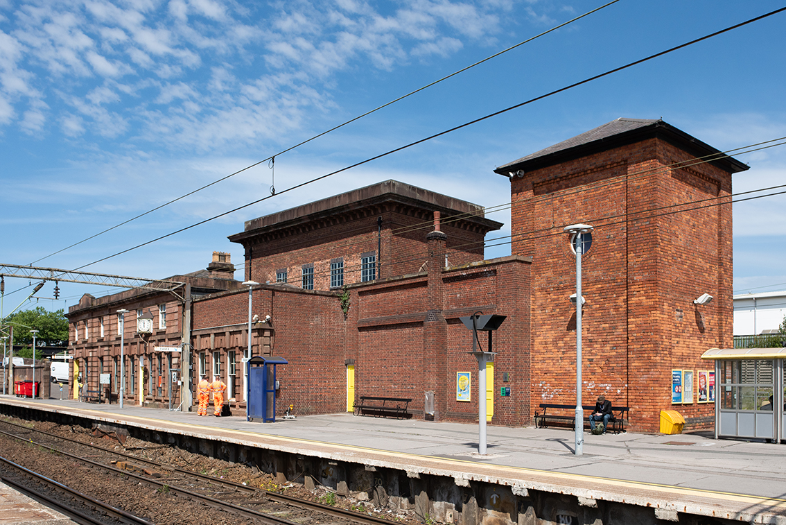 View of the brick-built hydraulic plant house and tall accumulator tower at Edge Hill Station, standing beside the platform with historic station buildings stretching behind.