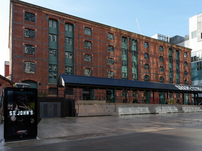 Wide view of the large brick warehouse building with rows of windows and green hoist doors, now part of the modern Grape Street development.