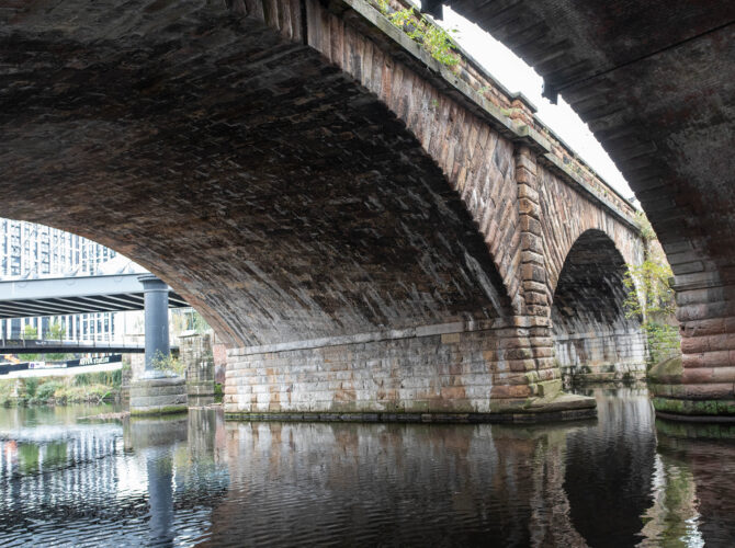 A close view from the riverbank beneath the stone arches of the 1830 railway bridge, showing the masonry and structure supporting the Liverpool and Manchester Railway.
