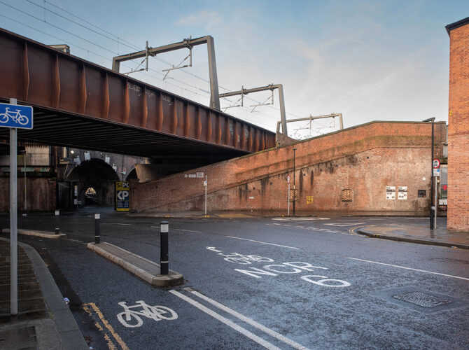 Angled view of the long brick ramp leading up towards the railway, set below the bridge and bordered by the street.