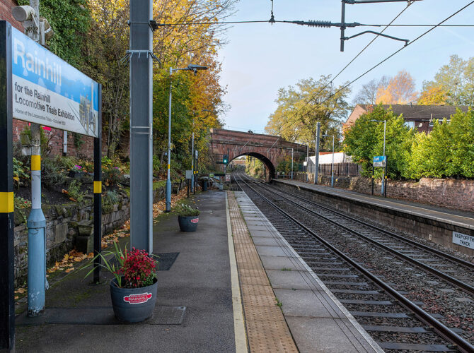 Platform view with Rainhill sign in the foreground and Skew bridge in the distance framed by autumn trees.