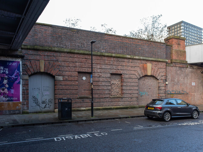 Street view of the historic station wall featuring a sealed arched entrance, smaller blocked windows, and layered brick detailing along Water Street.
