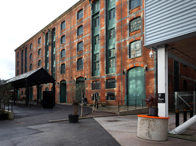 Angled view showing the warehouse façade with arched ground-floor openings and upper loading doors, alongside newer surrounding structures.