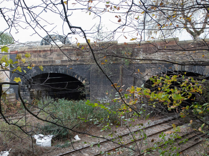 Two brick arches of Eccles railway bridge seen through trees and vegetation, carrying the railway over the tracks below.