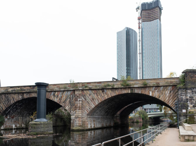 The historic Liverpool and Manchester Railway bridge spanning the River Irwell, with modern skyscrapers rising in the background.