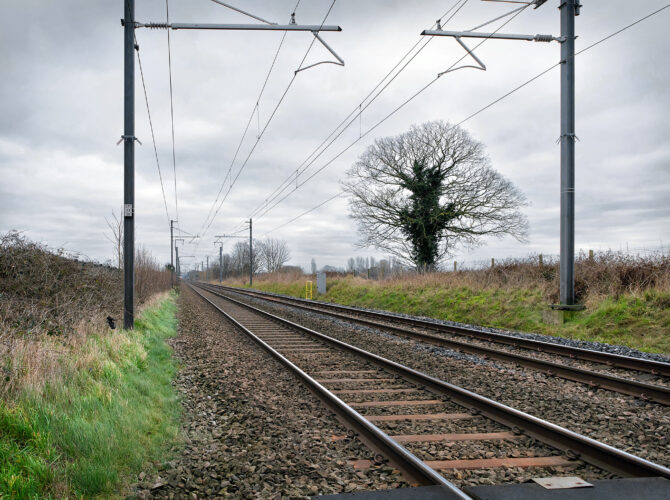 Railway tracks running straight across Chat Moss with overhead wires and a lone tree nearby.