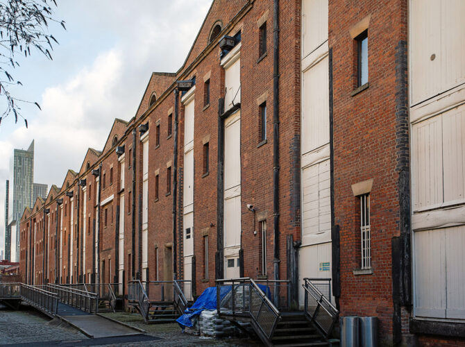 Angled view along the line of historic brick warehouses, featuring repetitive bays, loading doors, and external steps facing the former goods yard.