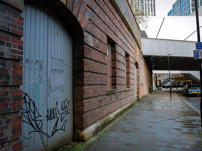 Angled view highlighting a boarded-up arched doorway with graffiti, set within the original brickwork of the 1837 Arrivals Station.