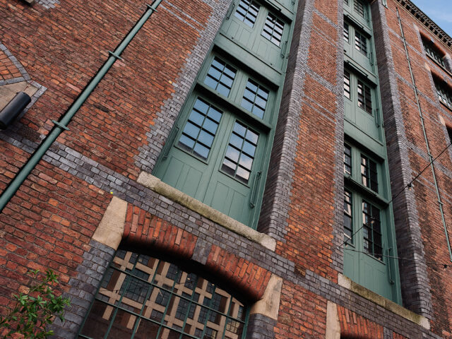 Upward view of the warehouse’s red brick wall with tall green loading doors and windows set between vertical brick piers.