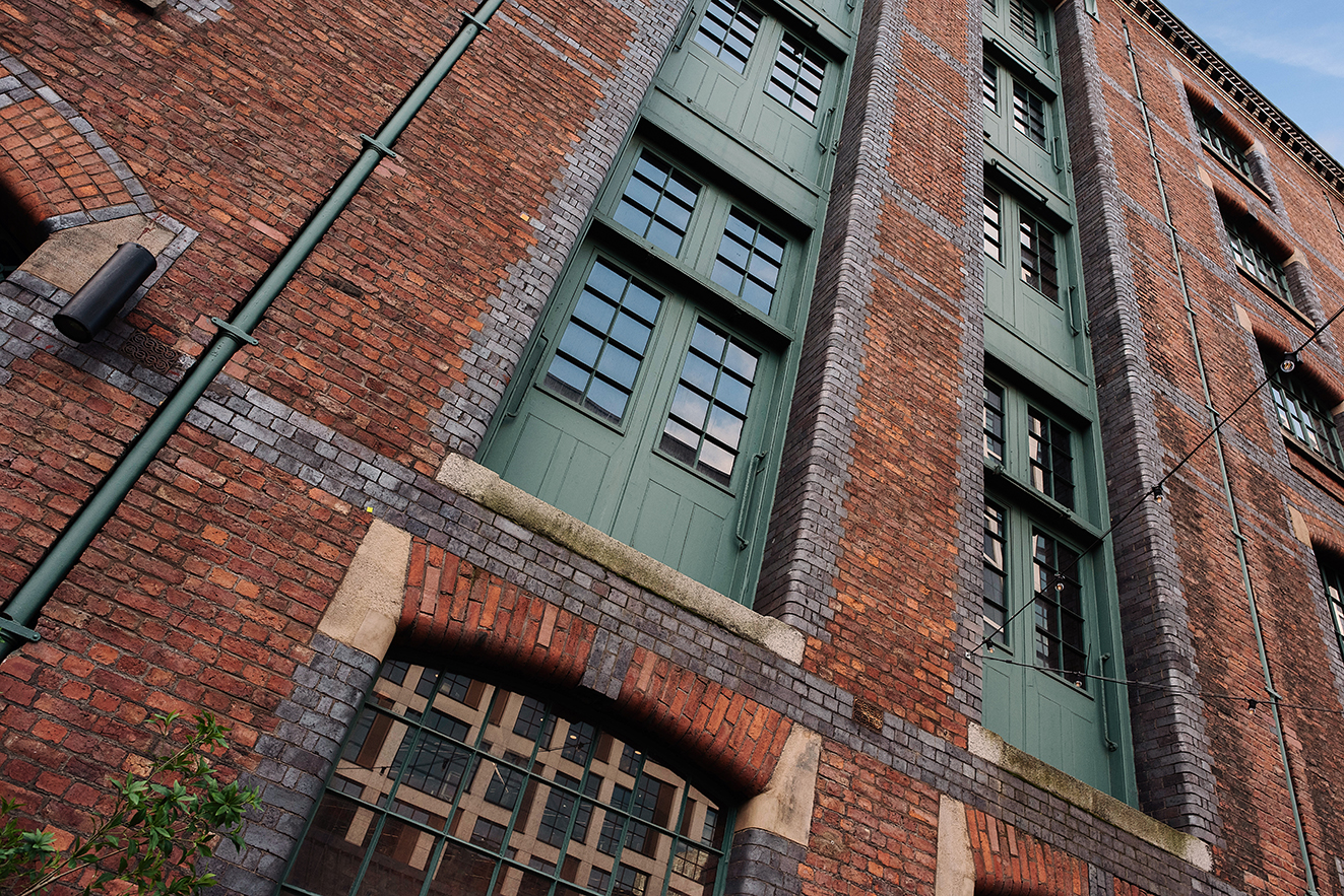 Upward view of the warehouse’s red brick wall with tall green loading doors and windows set between vertical brick piers.