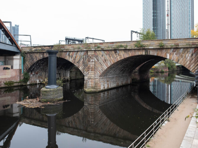 A clear view of Stephenson’s two-arch sandstone railway bridge crossing the River Irwell, with modern Manchester buildings and riverside paths nearby.