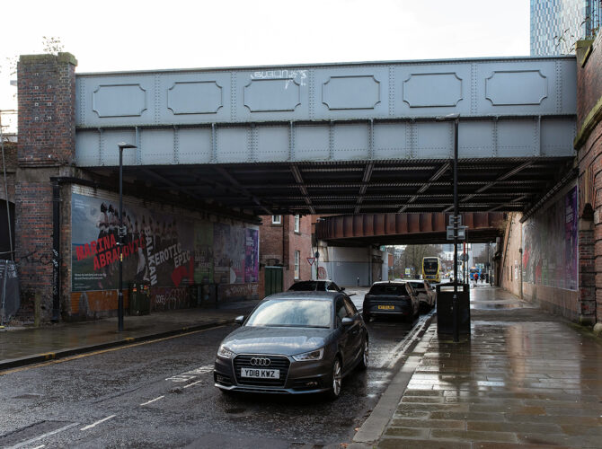 Street-level view beneath the railway bridge with vehicles parked along the road and brick arches visible to one side.