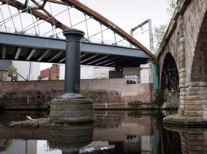 Close view of the cast-iron column rising from the river on a stone plinth, positioned beside the viaduct and beneath later bridges, indicating the support for a lost railway span.