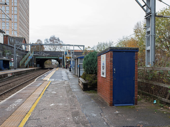 View along the platform at Eccles station showing the electrified tracks, the railway bridge ahead and a small brick lineside building with a blue door beside the platform.