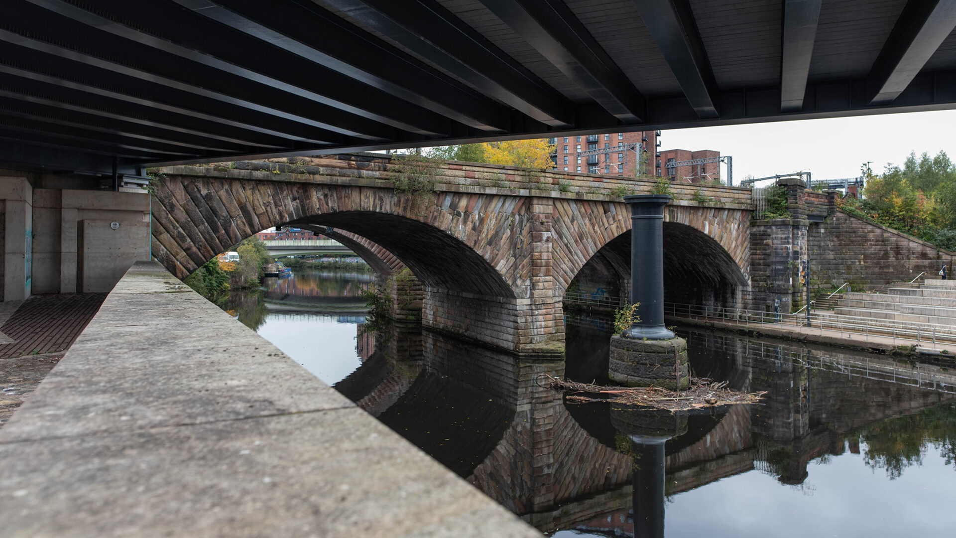 The 1830 sandstone railway bridge over the River Irwell seen from beneath a modern road bridge, showing its two stone arches reflected in the water.