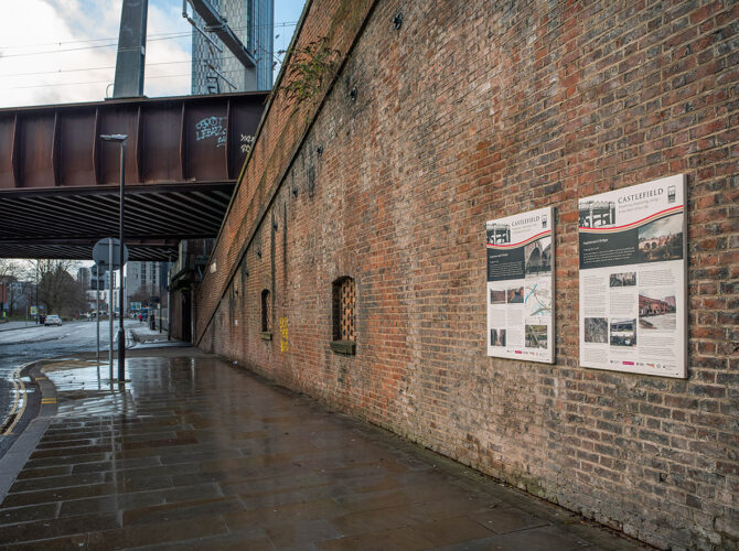 View along a brick wall beneath the railway showing information panels explaining the history of the linking bridge and surrounding area.