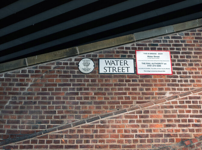 Close-up of street and railway signage mounted on the brick wall beneath the bridge, identifying the location as Water Street.
