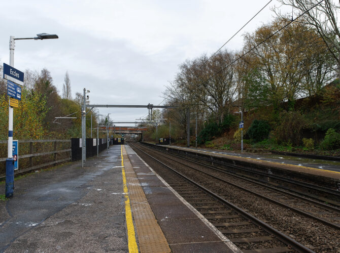 Wide view along the platforms and railway tracks at Eccles station with electrification gantries and station signage visible.