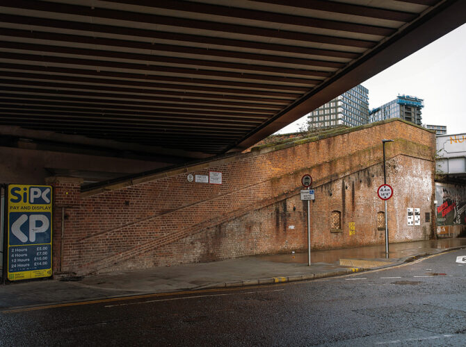 View of the brick cattle ramp rising alongside the road beneath the steel railway bridge, showing its original function linking to the station above.