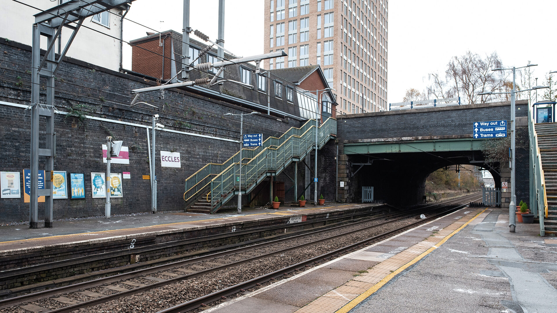 Platforms at Eccles station with the metal footbridge and brick railway bridge spanning the tracks, with station signage and surrounding buildings visible.
