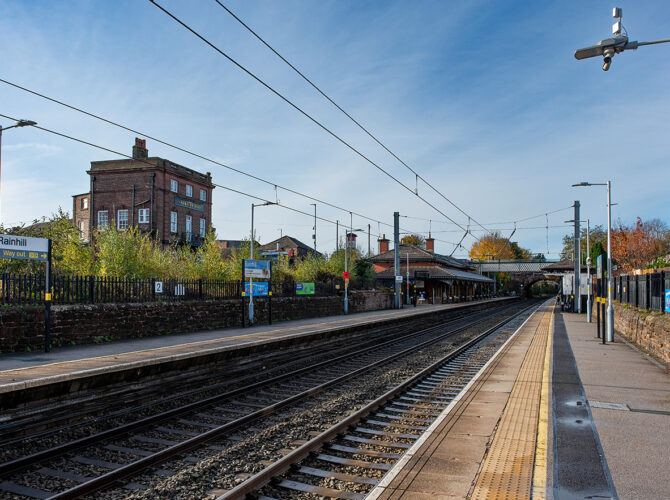 Wide view of the station with tracks, platforms, signage, and nearby houses under clear skies.