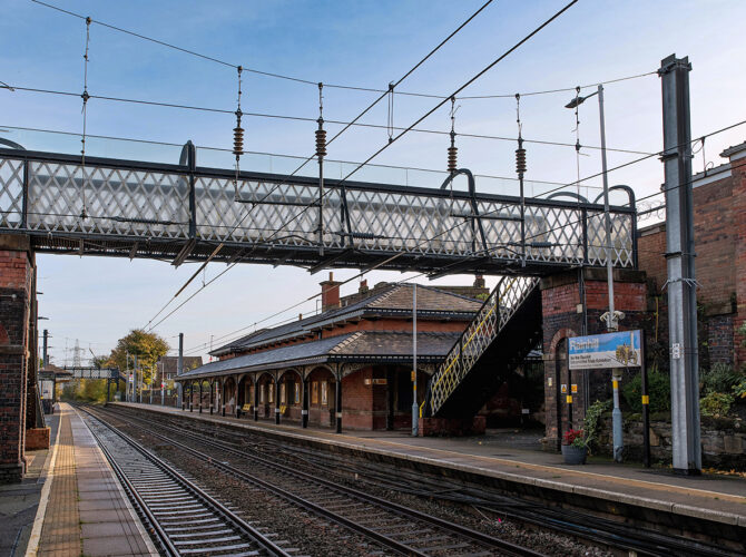 Wide view of Rainhill station showing platforms, a lattice footbridge, and traditional brick station buildings.