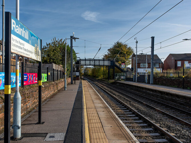 Long view down the platforms with a footbridge ahead, surrounded by autumn foliage and residential buildings.