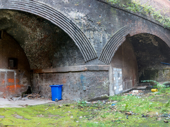 View of a large brick arch leading into the goods station, set within a walled cutting with moss, weeds, and scattered debris across the ground.