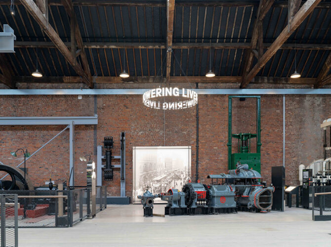 Wide interior view showing large stationary steam engines and industrial machines displayed along a central walkway beneath a timber roof.