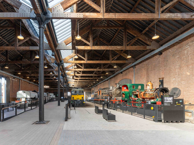 Interior scene featuring railway vehicles and industrial machines under the open trussed roof of the historic building.