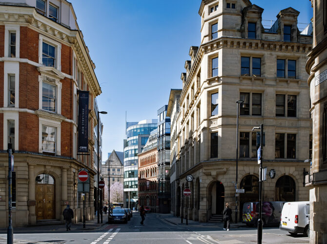Street scene at the junction of Fountain Street and Spring Gardens, showing a mix of ornate historic buildings and newer glass-fronted offices, with pedestrians and vehicles.