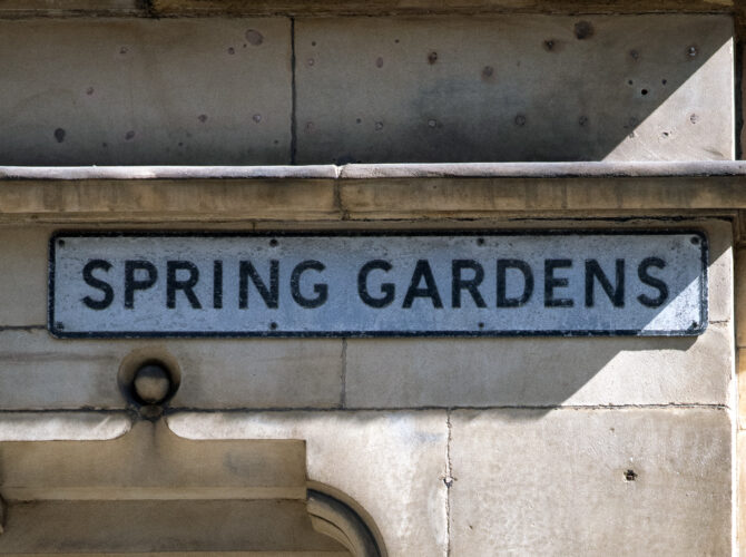 Close-up of a traditional street sign set into a stone façade, showing the name Spring Gardens in black lettering on a white background.