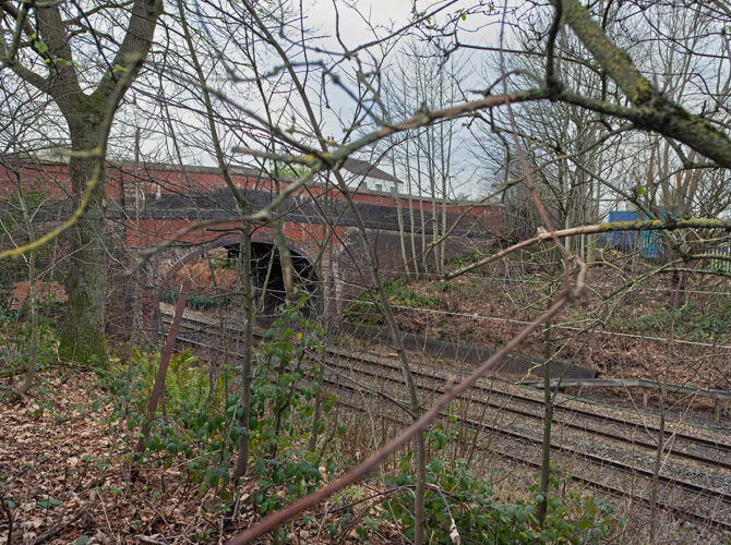 Seen through branches and foliage, the red-brick arch of Broseley Bridge spans the railway cutting below. The structure is partially hidden, highlighting how the bridge sits within the landscape.