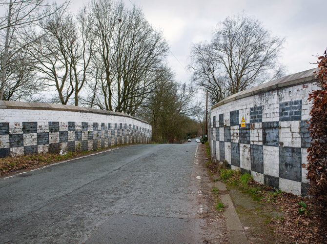 A road crosses Broseley Bridge between distinctive black-and-white chequered parapet walls. The curved walls guide the road gently over the railway, with safety signage visible along the route.