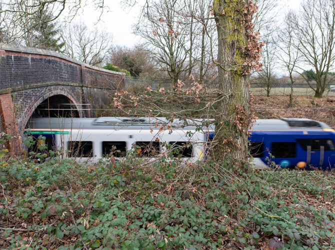 A passenger train speeds beneath Broseley Bridge, partially blurred in motion. The brick arch frames the track below, while trees and undergrowth surround the cutting, emphasising the bridge’s long-standing role over a busy line.
