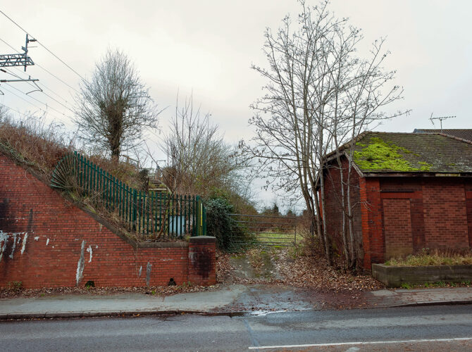The former station site, now an empty roadside space with a brick embankment, metal fencing and steps leading up towards the railway line.