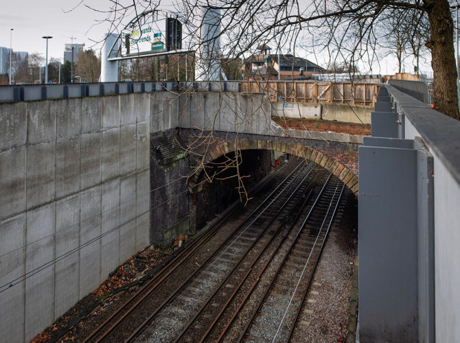 Angled view of the brick arch bridge with tracks below, framed by modern concrete retaining walls and roadside structures