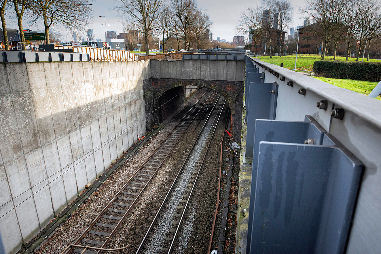 View along the railway line running through a concrete-lined cutting beneath an older brick arch bridge