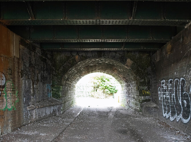Tunnel-like arch with brick walls and iron beams, leading to light beyond.