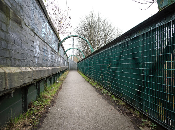 Narrow pedestrian path with metal fencing and green iron supports along the bridge.
