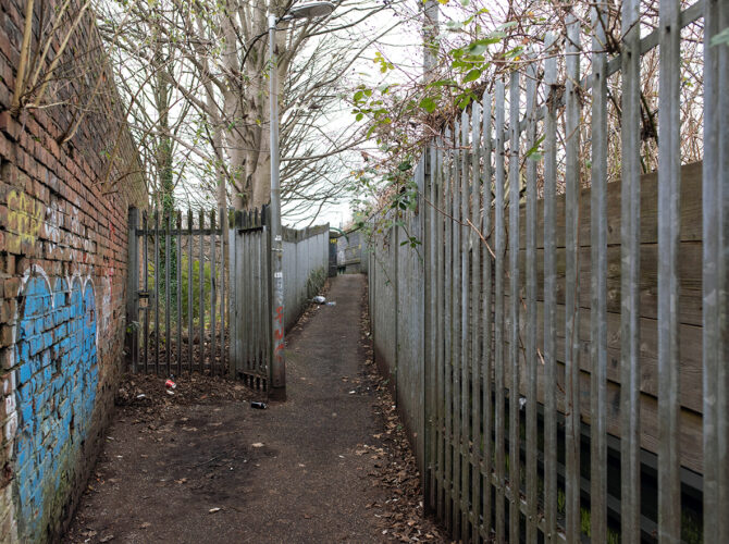 Enclosed path between brick wall and fencing approaching the bridge area.