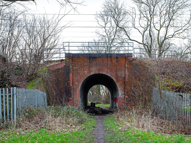 A head-on view of the brick accommodation bridge, framed by trees and fencing, with a worn footpath leading to the arch.
