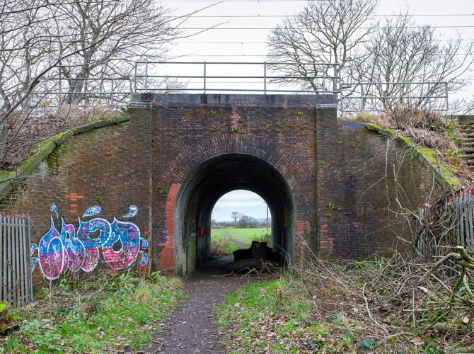 The bridge from closer range, showing the arched entrance, brick detailing and graffiti, with fields visible beyond.