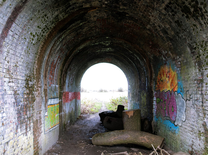 Inside the tunnel, the curved brick roof and damp walls create a dark passage leading out to daylight.