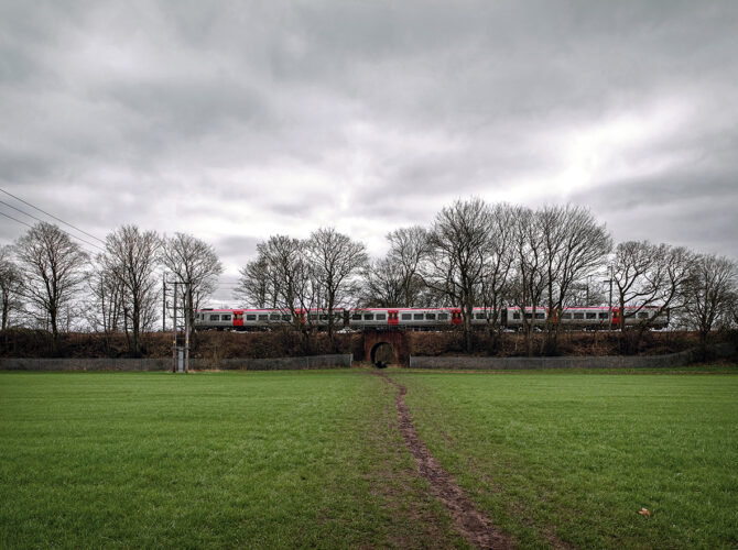 A distant landscape view of the bridge beneath the railway, with a modern train passing overhead across open fields.