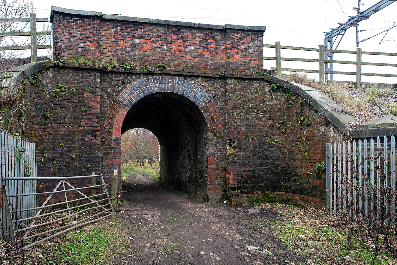 A wider approach to the bridge showing rural fencing and a gate leading towards the tunnel.