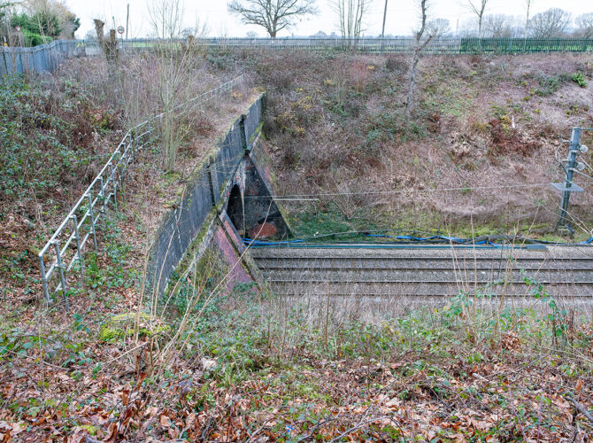 A front view of the main tunnel portal, with a wide brick arch beneath a road bridge and railway tracks leading directly into the dark opening.