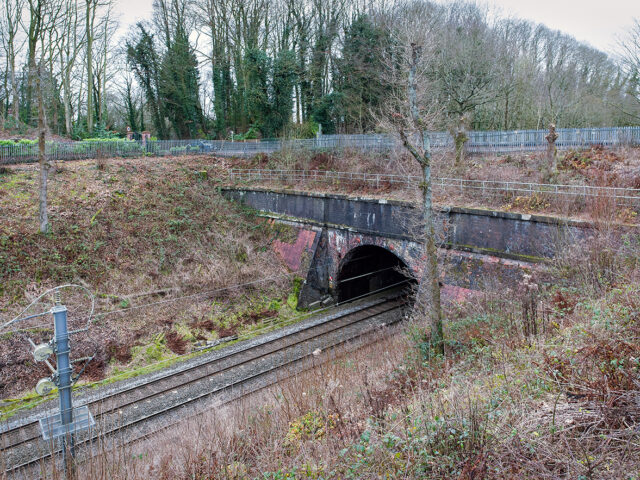 A side view of Kenyon Tunnel showing a smaller brick opening tucked into the cutting wall, with railway tracks and cables running past in the foreground