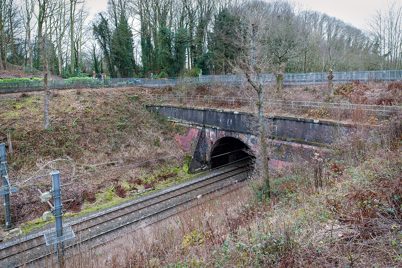A side view of Kenyon Tunnel showing a smaller brick opening tucked into the cutting wall, with railway tracks and cables running past in the foreground