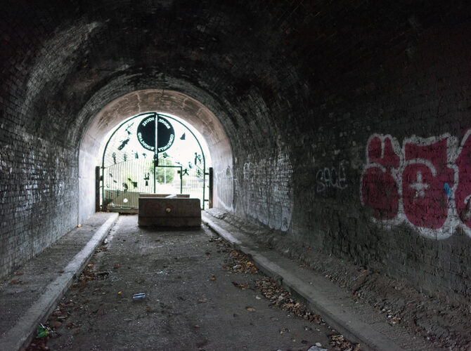 The curved brick-lined tunnel stretches ahead, with light filtering in from the gated opening at the far end.